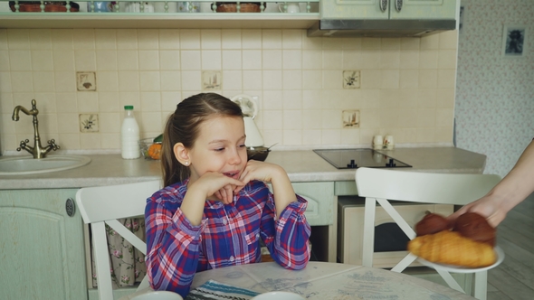 Young Mother Bringing Tasty Muffins and Croissants To Her Cute Daughter Sitting at the Table in