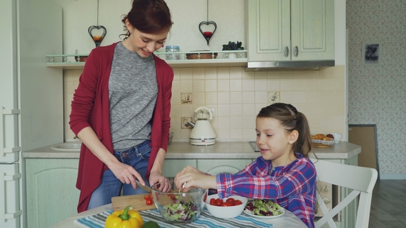 Happy Young Mother and Cheerful Daughter Cook Salad Together in the Kitchen at Home Cutting
