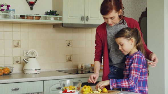 Young Pretty Mother Teaching Her Cute Daughter To Cut Vegetables Properly. Little Girl Cooking
