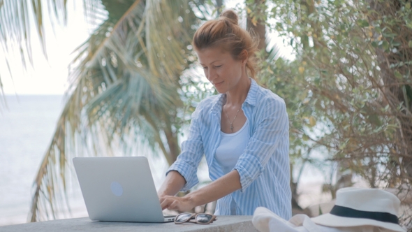 Young Woman Sitting at the Table with a Laptop with Sea View Behind
