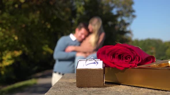 Closeup on a Wedding Ring, a Present and a Red Rose in a Park - a Happy Couple Hugs in Background