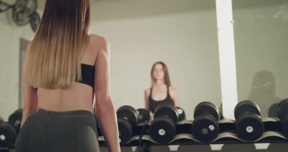 Rear View of Sports Girl Reflected in a Gym's Mirror Does Squats with Dumbbells