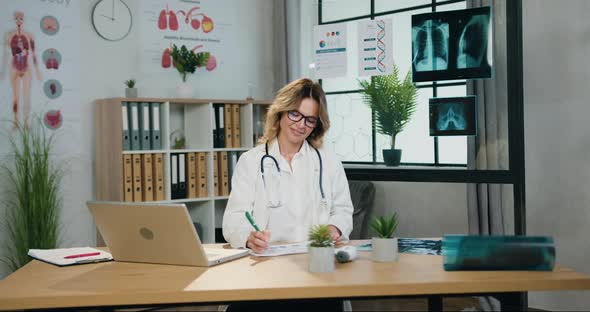Female Doctor in Glasses and Hospital Uniform Working with Paper Reports in Medical Office