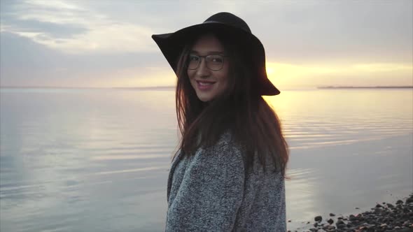 Young Pretty Woman in Black Hat and Glasses Near the Sea at Sunset