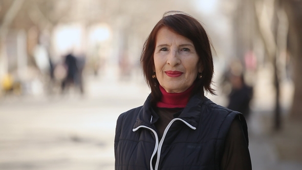 An Optimistic Woman in a Stylish Jacket Smiles and Stands in the Sunny Alley in Spring