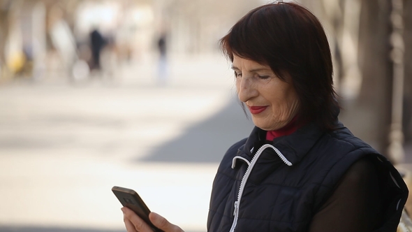 A Smart Woman Surfs the Net in Her Smartphone and Sits on a Bench in Spring