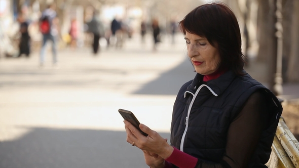 A Slender Woman Browsing the Net in Her Smartphone and Sits on a Bench in Spring