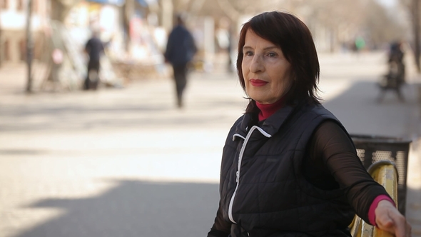 A Middle-aged Woman in a Sportive Jacket Sits on a Bench and Relaxes in Spring