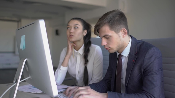 Two Young Colleagues Are Talking, in Work Process, Sitting at Table with Computer in Modern Office