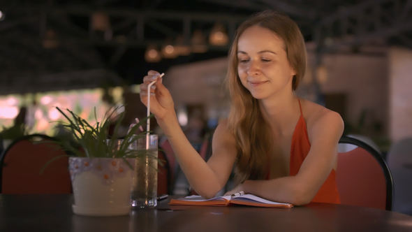 Girl Stirs Cocktail with Straw at Table Smiles