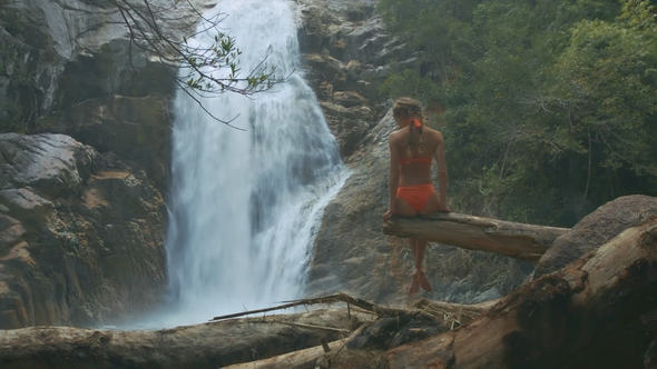 Slim Woman Sits on Tree Takes Pleasure in Waterfall