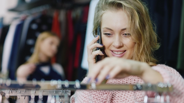 Portrait of a Young Woman Chooses a Dress in the Store, Talking on a Cell Phone.