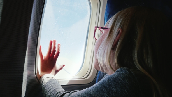 A 6-Year-old Girl with Spectacles Sits in a Plane, Looks Out the Window. Her Hand Is Pressed To the