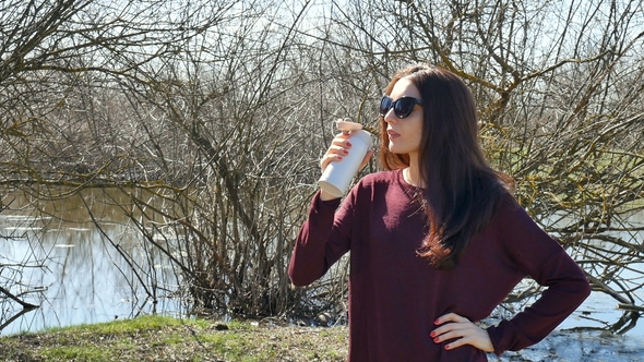 Young Brunette Woman in Sunglasses Drinking Some Beverage Using Thermo Cup on Lake Background