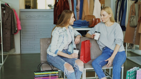 Two Charming Girls Sitting in Clothes Shop with Coffee, Talking and Laughing After Shopping