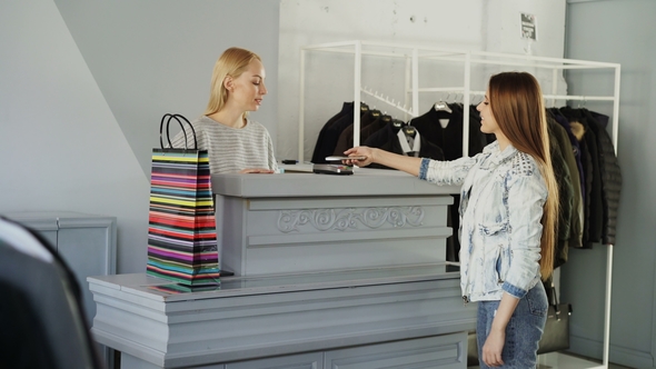 Cheerful Customer Is Using Smartphone To Pay for Purchased Clothes While Standing at Cashier's Desk