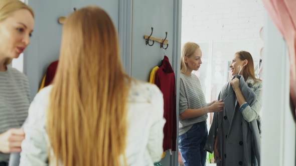 Pretty Young Woman Is Checking Fashionable Coat in Fitting Room with Her Friend Helping Her To