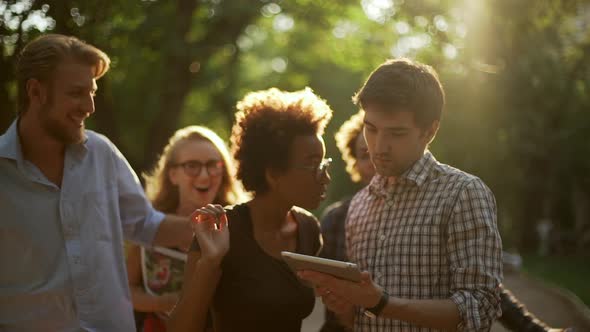 African American Female Standing in Park with Caucasian Male with Tablet Talking While Their Friends