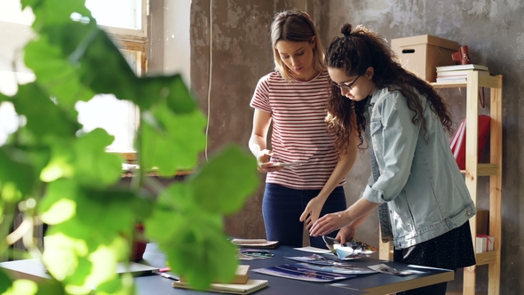 Young Women Are Standing Near Table, Placing Pictures on It and Shooting Flat Lay with Smartphone