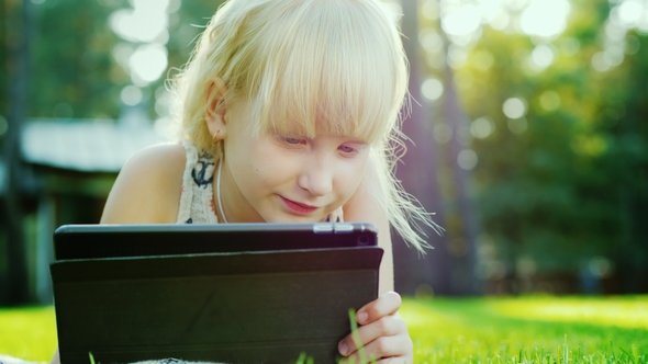 Blonde Girl 6 Years Playing on the Tablet, Emotionally Reacts. Lying on the Lawn in the Backyard of