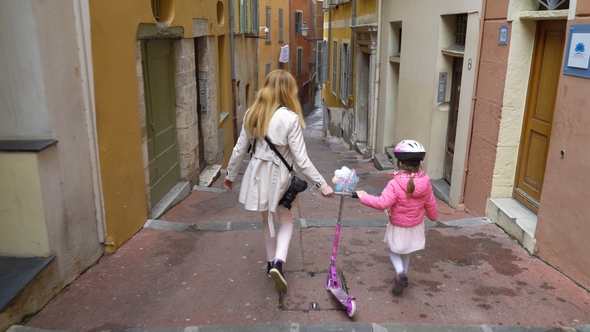 Mother and Her Daughter Walk along the Narrow Street of the Old European City with Scooter