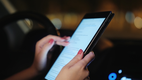 A Woman Uses a Tablet Inside the Car. Evening, the Traffic of Cars Is Visible Outside the Window. In