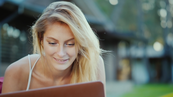 Beautiful Young Woman Enjoys a Laptop. Lies on the Lawn in the Backyard of the House in the Sun at