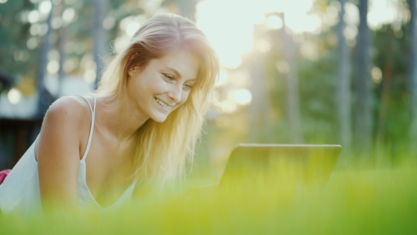 Beautiful Young Woman Enjoys a Laptop. Lies on the Lawn in the Backyard of the House in the Sun at