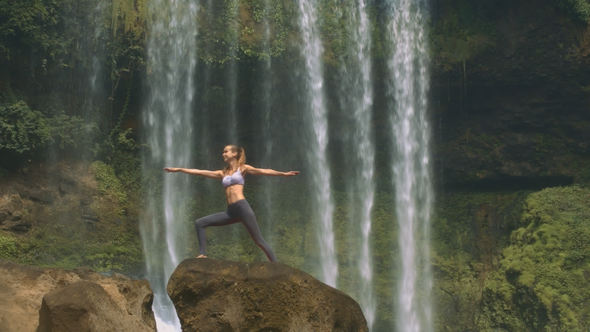 Young Woman Does Yoga on Rock against Waterfall