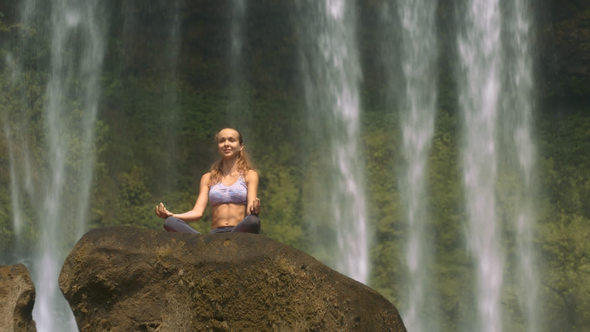Girl Sits in Lotus Pose on Brown Rock by Waterfall