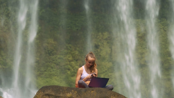 Girl Sits with Laptop and Phone on Cliff at Water