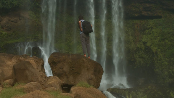 Guy with Backpack Standing on Rock Drinks Water by Waterfall