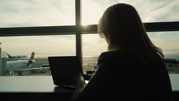 A Woman Is Using a Laptop at the Airport Outside the Window You Can See Planes