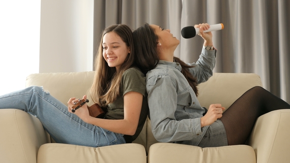Two Sisters Having Fun on the Couch, One Is Playing at Ukulele and the Other Is Singing at a