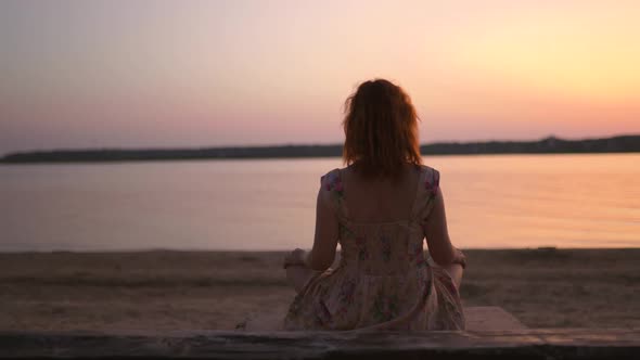 Young Woman Meditating at River During Sunset with Boat Passing By