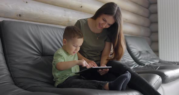 Happy Mom and Son Play on a Digital Tablet in a Spacious Living Room on the Sofa