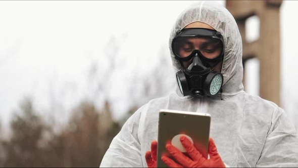 Man in Bio-hazard Suit and Gas Mask Takes Notes in His Tablet Standing on the Polluted Land