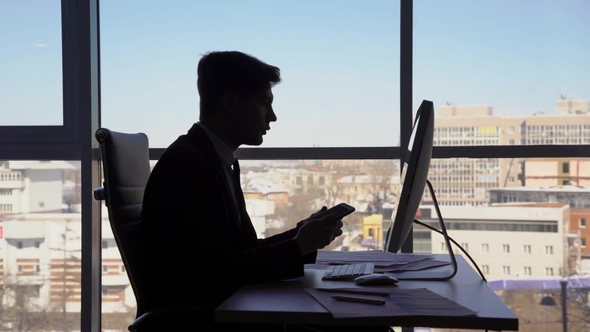 Young Handsome Businessman Is Working, Sitting at Table, Talking on Phone on Background of Window in