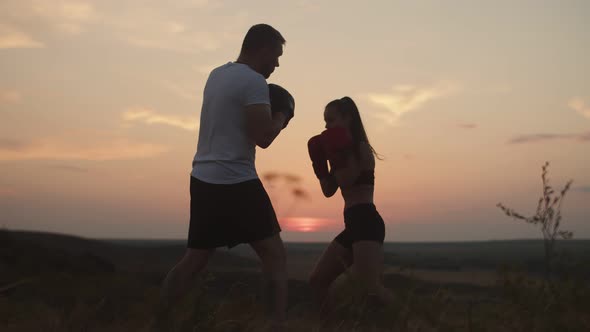 Woman Boxing Gloves Trains with Man Paws of Boxing in Nature Against the Background of the Sunset