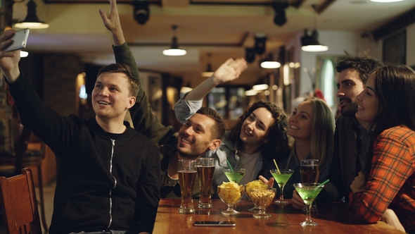 Young Blogger Is Recording Video with Smartphone Sitting at Table in Bar with His Friends