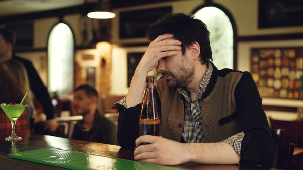 Upset Unshaven Guy Is Sitting at Bar Counter with Bottle of Beer, Sighing and Touching His Face in