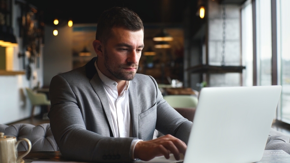 a Young Businessman in a Gray Stylish Suit Works at a Laptop in a Cafe Near the Window