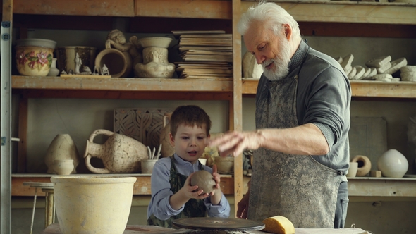 Caring Silver-haired Grandfather Is Teaching Young Cute Grandson To Work with Clay on Throwing-wheel