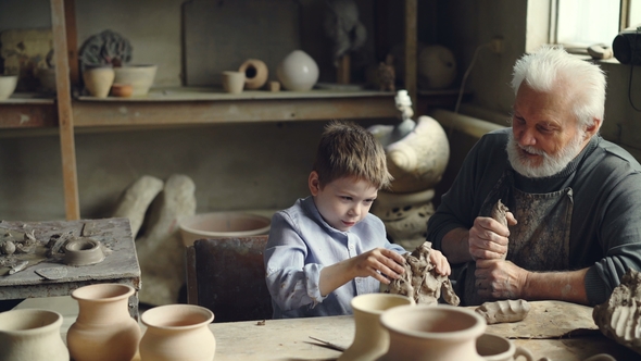 Cute Boy Is Having Fun Playing with Clay in His Grandfather's Pottery Studio While Elderly Man 