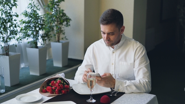 Well-dressed Angry Young Man Is Waiting for His Girlfriend in Restaurant, Using Smartphone