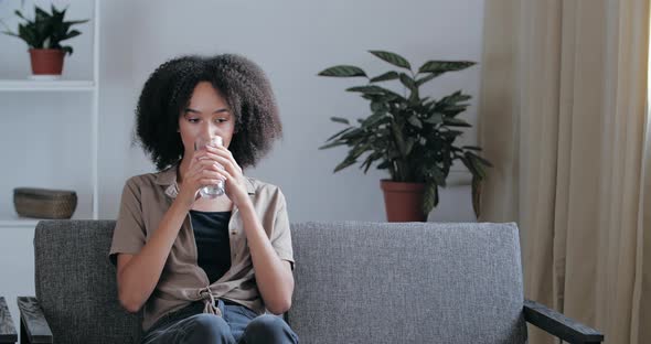 Calm Smiling Dreamy African Young Woman Sit on Sofa Drinking Pure Still Cool Water Relaxing at Home