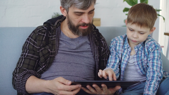 Loving Father and His Cute Little Child Are Using Table Touching Screen and Talking While Sitting 