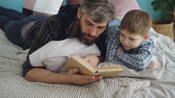 Loving Parent Bearded Man Is Reading Interesting Book To His Curious Little Son on Double Bed