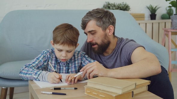 Child Is Learning, Writing Words in Exercise Book and His Father Is Teaching Him Sitting Near Table