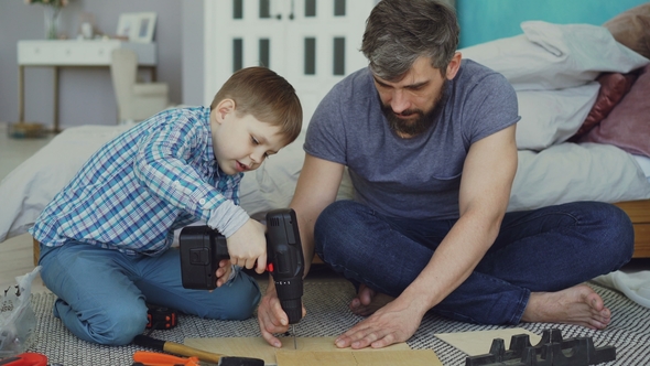 Little Boy Is Learning To Use Electric Screwdriver While His Dad Is Explaining How To Work